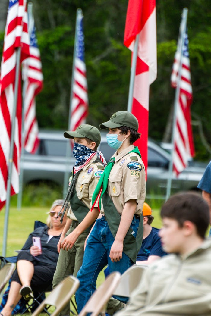 Two scouts in uniforms and masks walk outdoors during a flag ceremony with spectators.