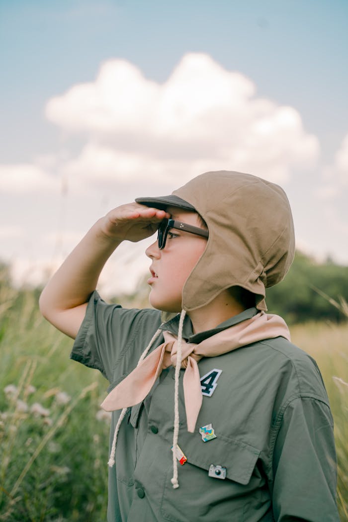 A young scout in uniform shielding his eyes while looking ahead in a green field under a bright sky.