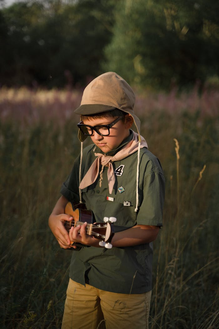 A young boy scout with eyeglasses and hat plays a ukulele outdoors in a natural setting.