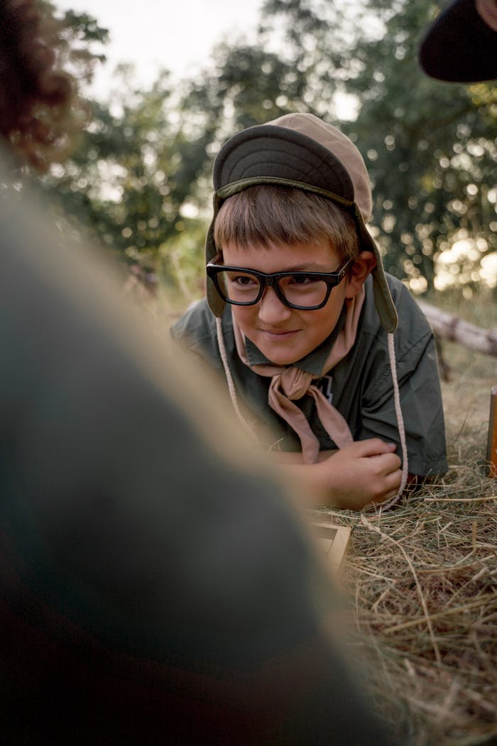 A young boy scout wearing eyeglasses and a hat lies in the grass during a summer camp outdoors.