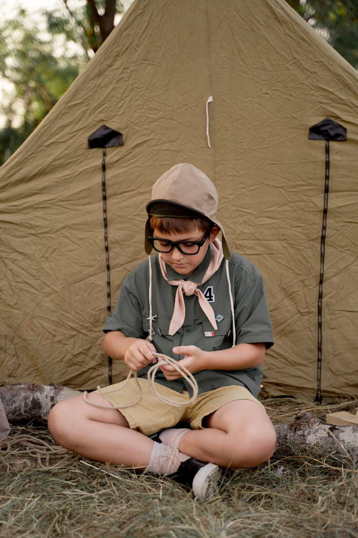 A young scout wearing eyeglasses and a uniform sits by a tent, focusing on a rope task during a camping trip.