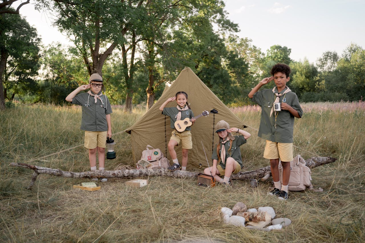 Group of diverse kids at a summer scouting camp, saluting in front of a tent in a forest.